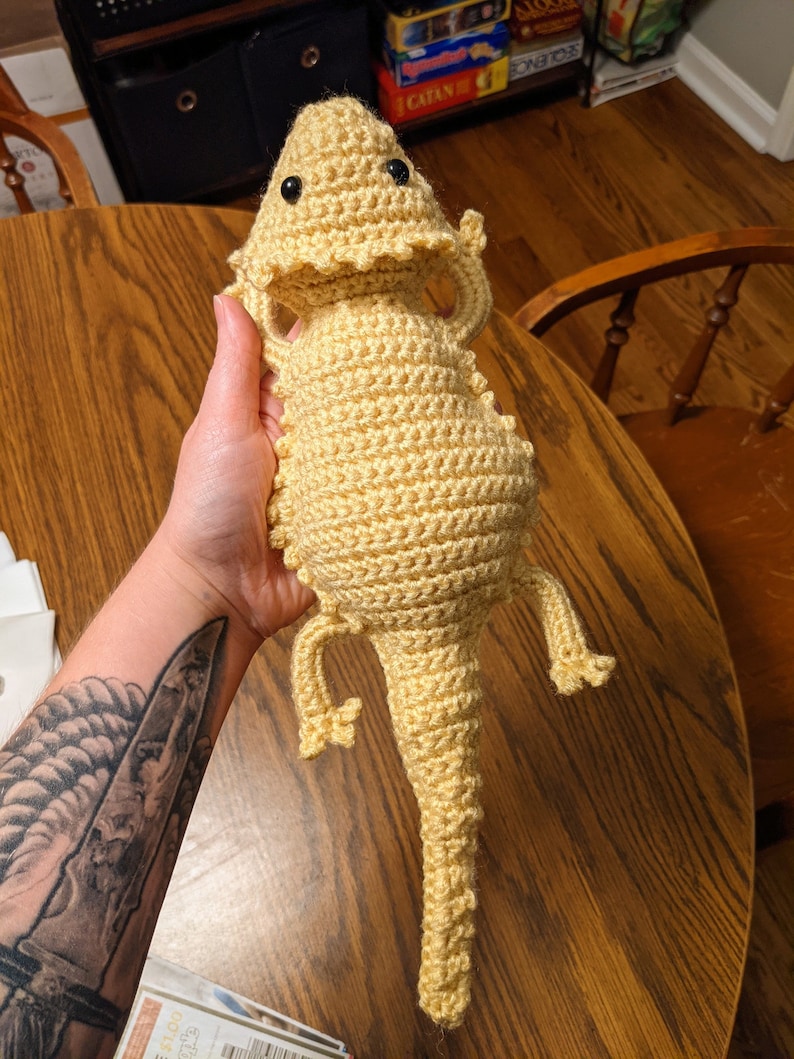 May include: A crocheted yellow bearded dragon plush toy with black eyes. The toy is being held by a hand with a tattoo.