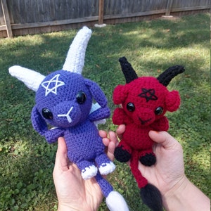 Op de afbeelding: Twee gehaakte pluche speelgoedjes, een paarse met witte hoorns en een witte staart, de andere rood met zwarte hoorns en een zwarte staart. Beide speelgoedjes hebben een pentagram op hun borst.