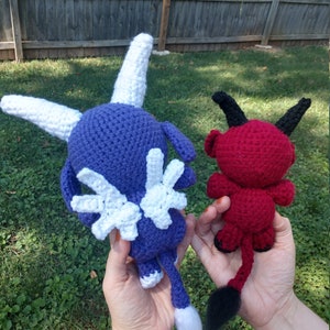 Op de afbeelding: Twee gehaakte knuffels, een paarse met witte vleugels en een witte staart, de andere rood met zwarte hoorns en een zwarte staart.