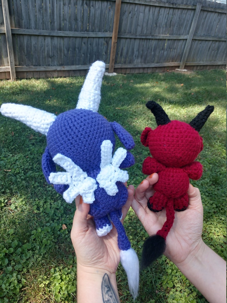 Op de afbeelding: Twee gehaakte knuffels, een paarse met witte vleugels en een witte staart, de andere rood met zwarte hoorns en een zwarte staart.
