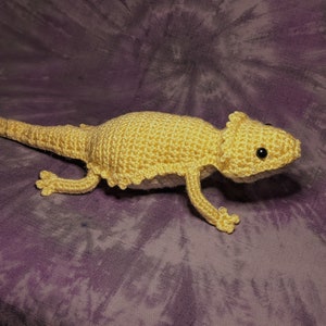 May include: A crocheted yellow bearded dragon plush toy with black eyes. The toy is lying on a purple and white tie-dye fabric.