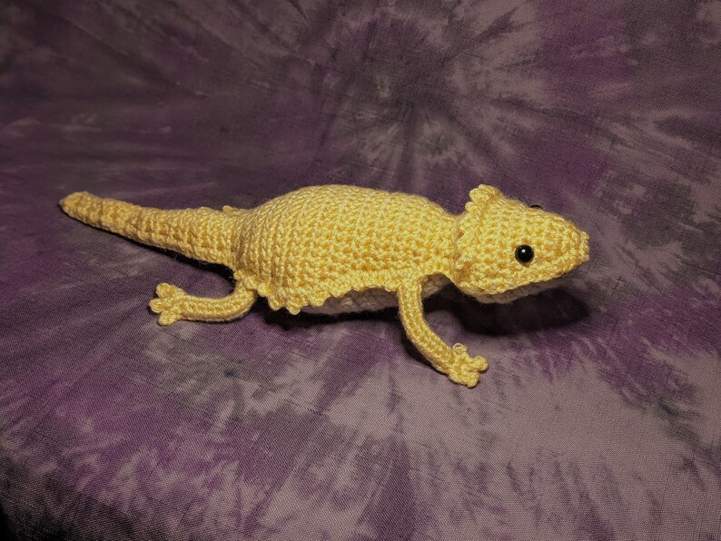 May include: A crocheted yellow bearded dragon plush toy with black eyes. The toy is lying on a purple and white tie-dye fabric.