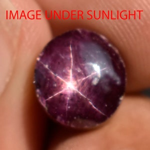 Puede incluir: Primer plano de una gema de color púrpura oscuro con un patrón de estrella de seis puntas. La gema tiene forma ovalada y parece estar bajo la luz del sol. El texto "IMAGE UNDER SUNLIGHT" es visible en rojo.