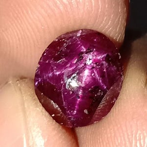 May include: A close-up of a faceted, oval-shaped ruby gemstone. The ruby is a deep, rich magenta color with internal inclusions and a reflective surface. The gem is held between two fingers.