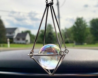 Puede incluir: Un amuleto de coche plateado con una esfera de vidrio iridiscente que cuelga de una cadena. El amuleto tiene la forma de una jaula geométrica.