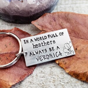 Pu&ograve; includere: Un portachiavi d'argento con un'etichetta rettangolare su cui &egrave; scritto "In a world full of heathers always be a Veronica". L'etichetta ha un piccolo disegno di fiore sul lato destro.