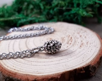 May include: A silver necklace with a pine cone pendant. The necklace is on a wooden surface.