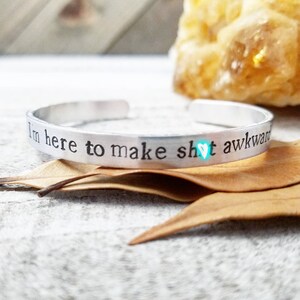 May include: A silver cuff bracelet with the stamped text "I'm here to make sh!t awkward" and a small turquoise heart symbol. The bracelet is displayed on a white and gray surface with dried leaves.