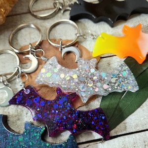 Pode incluir: Cinco chaveiros diferentes em forma de morcego com glitter e um charme de lua crescente. Os chaveiros s&atilde;o feitos de resina e v&ecirc;m em uma variedade de cores, incluindo preto, laranja, branco, prata e roxo.