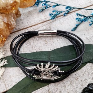 May include: A black leather bracelet with a silver charm featuring a pentagram inside a circle with rays emanating from the circle. The bracelet has a magnetic clasp.