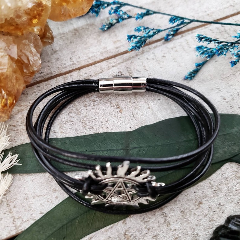 May include: A black leather bracelet with a silver charm featuring a pentagram inside a circle with rays emanating from the circle. The bracelet has a magnetic clasp.