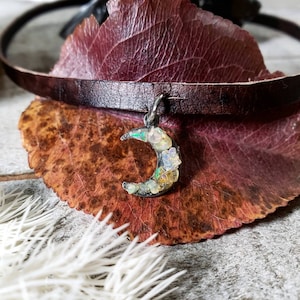 May include: A leather choker necklace with a crescent moon charm. The charm is filled with iridescent opal chips.