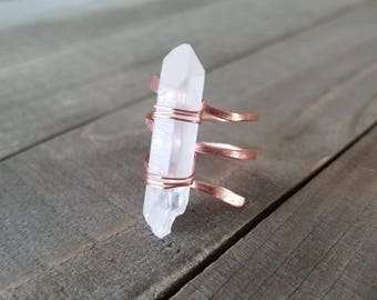 May include: A clear quartz crystal wrapped in copper wire, forming a ring. The ring is on a wooden surface.