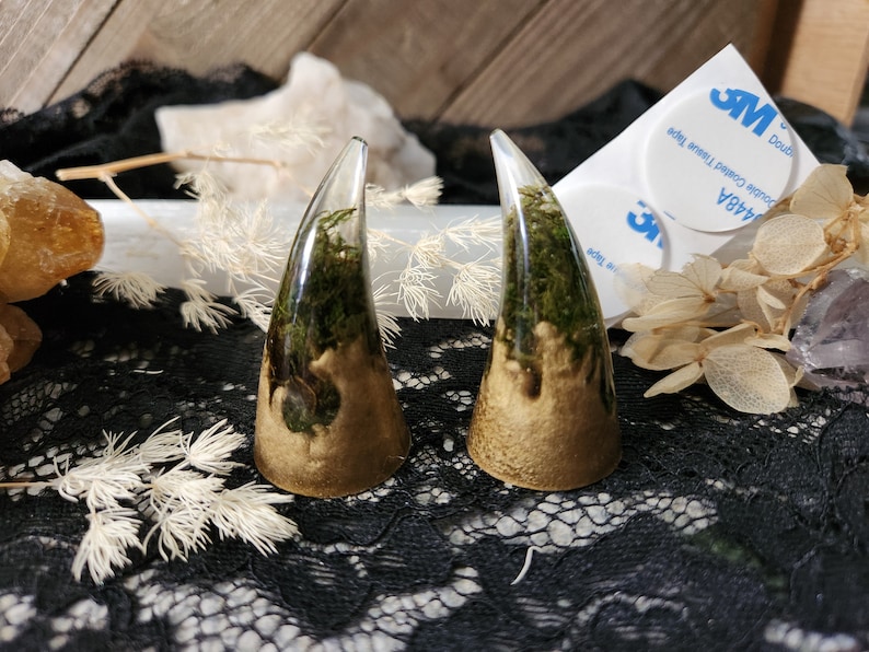 May include: Two resin horns with a gold base and green moss inside. The horns are on a black lace surface.