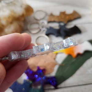 Pode incluir: Um chaveiro de morcego com glitter prateado com um anel de metal prateado. O chaveiro est&aacute; pendurado em uma m&atilde;o.