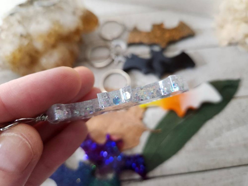 Pode incluir: Um chaveiro de morcego com glitter prateado com um anel de metal prateado. O chaveiro est&aacute; pendurado em uma m&atilde;o.