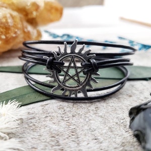 May include: A black leather bracelet with a silver pentagram charm. The charm is surrounded by a sunburst design.