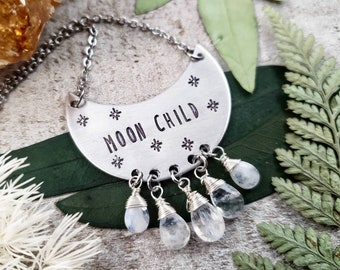May include: A silver crescent moon necklace with the words "Moon Child" inscribed on it. The necklace has five dangling clear quartz crystals.
