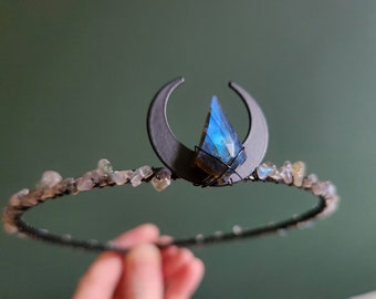 May include: A handcrafted crown featuring a crescent moon design and a faceted blue gemstone. The crown is adorned with small, irregular-shaped stones along the band. The metalwork is dark, and the overall aesthetic is mystical.