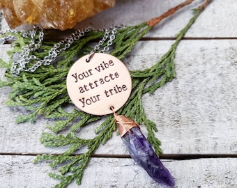 May include: A silver chain necklace with a round, light brown pendant. The pendant has the text "Your vibe attracts Your tribe" stamped on it. A purple crystal is wire-wrapped and hangs below the pendant. The necklace rests on green foliage.