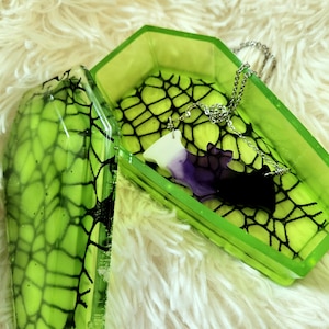 Può includere: Una scatola verde a forma di bara con un design a ragnatela nera. La scatola è aperta e all'interno c'è una collana a forma di pipistrello viola.