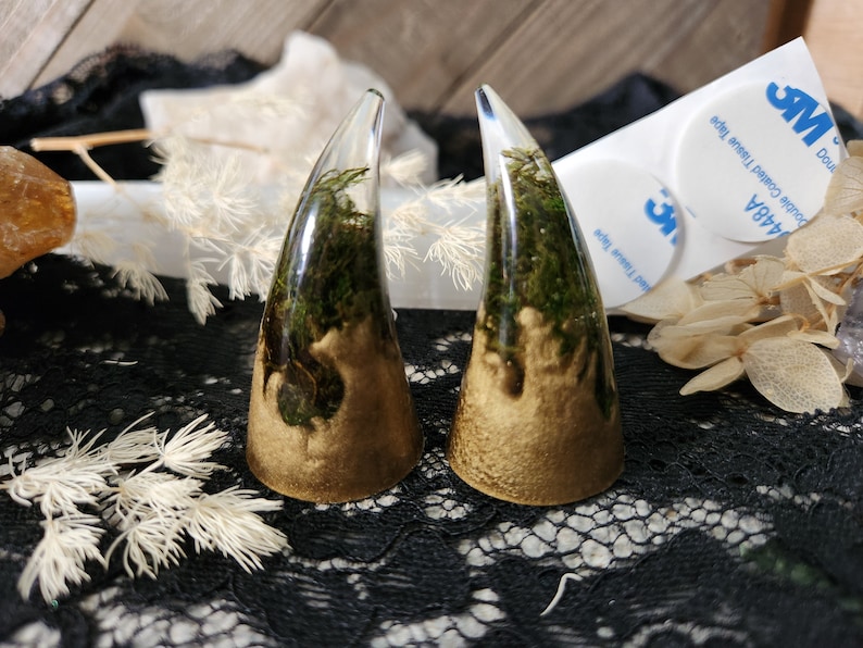 May include: Two clear resin horns with gold accents and green moss inside. The horns are sitting on a black lace surface.