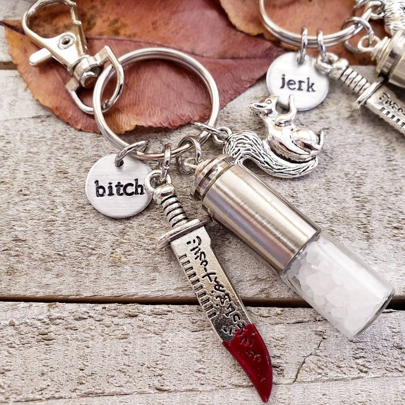 May include: A silver keychain with a silver charm of a squirrel, a silver charm with the word "bitch", a silver charm with the word "jerk", a silver charm of a knife with a red handle, and a silver charm of a small glass vial filled with white crystals.