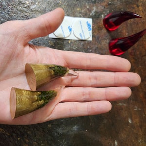 May include: Two pairs of resin horn plugs, one pair is a light brown with green moss embedded in the resin, the other pair is a clear red resin.