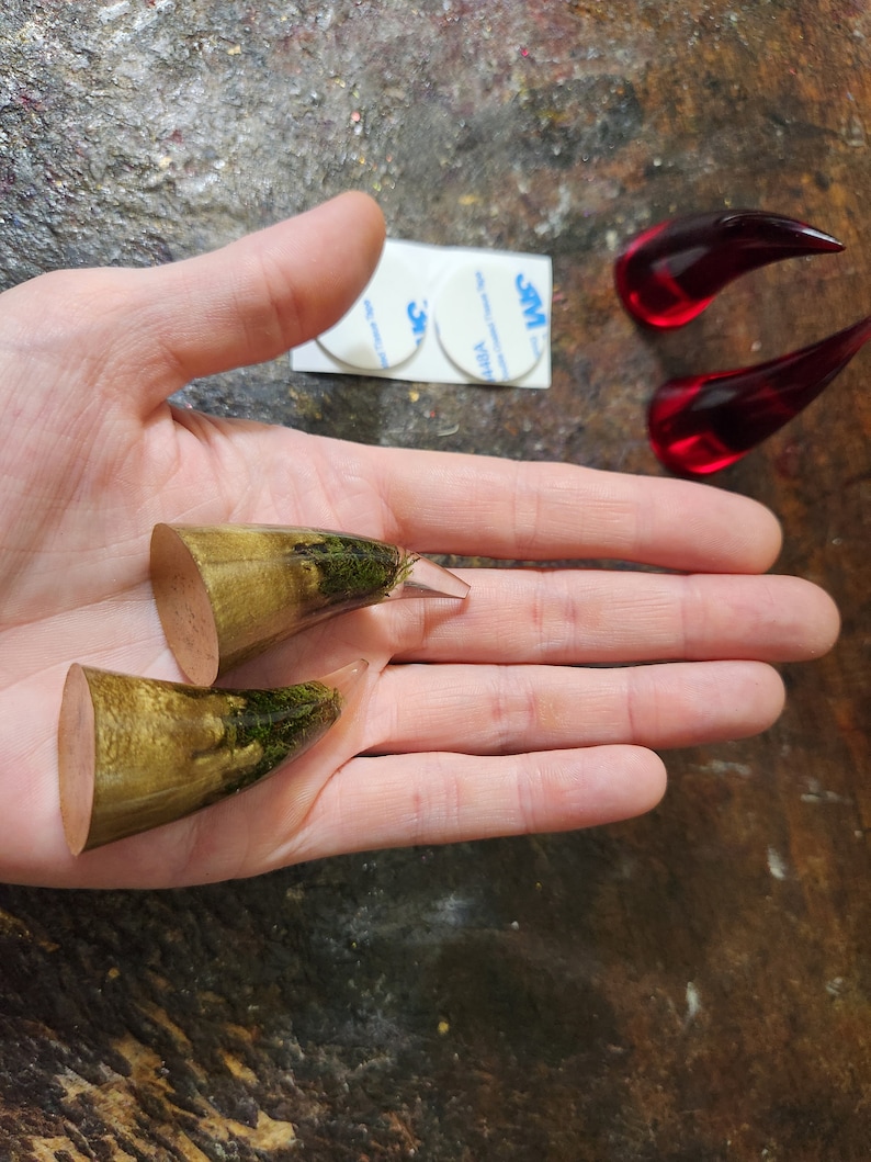 May include: Two pairs of resin horn plugs, one pair is a light brown with green moss embedded in the resin, the other pair is a clear red resin.