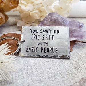 May include: A silver rectangular keyring with the quote "YOU CAN'T DO EPIC SHIT WITH BASIC PEOPLE" stamped in black. The keyring is attached to a silver ring. The background features crystals and dried leaves.