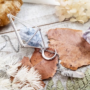 May include: A silver necklace with a geometric cage pendant containing a blue crystal. The pendant is attached to a silver crescent moon charm and a silver chain.