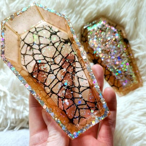 Op de afbeelding: Twee gouden glitterhars-sieradenbakken in de vorm van een kist. Een bak heeft een zwart spinnenwebontwerp erin.