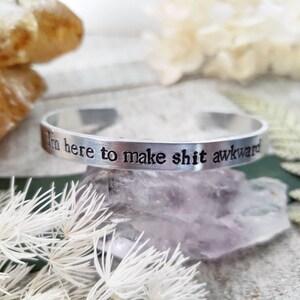 May include: A silver cuff bracelet with the phrase "I'm here to make shit awkward" engraved in black lettering. The bracelet is displayed on a purple crystal with white floral accents.
