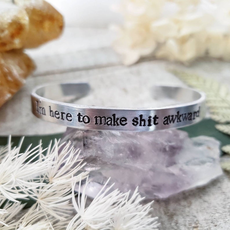May include: A silver cuff bracelet with the phrase "I'm here to make shit awkward" engraved in black lettering. The bracelet is displayed on a purple crystal with white floral accents.