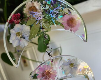 May include: A pair of crescent moon shaped earrings with pressed flowers and iridescent glitter embedded in clear resin. The earrings are connected by a silver chain.