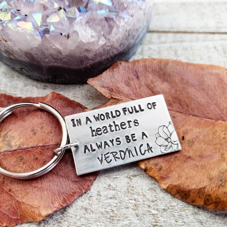 Pu&ograve; includere: Un portachiavi d'argento con un'etichetta rettangolare su cui &egrave; scritto "In a world full of heathers always be a Veronica". L'etichetta ha un piccolo disegno di fiore sul lato destro.