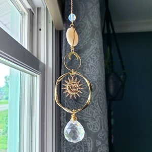 May include: A gold sun and moon crystal suncatcher with a clear crystal prism hanging from the bottom. The suncatcher is made of metal and has a beaded chain.