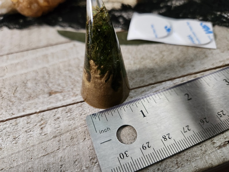 May include: A cone-shaped ring made of clear resin with gold and green moss inside. The ring is measured against a ruler, showing it is about 1 inch tall.