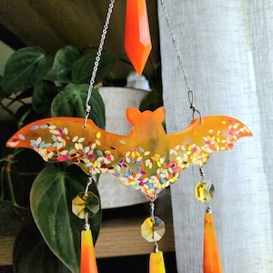 May include: Orange and glitter bat shaped resin suncatcher with crystal drops hanging from it.
