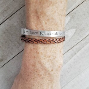 May include: Two bracelets are shown. The top bracelet is silver with the stamped text "I'm here to make shit awkward". The bottom bracelet is a braided brown leather cuff.