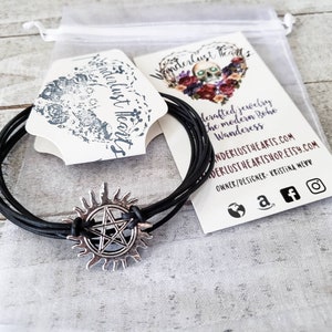 May include: Black leather bracelet with a silver charm featuring a pentagram inside a sunburst. The bracelet is packaged in a white organza bag with a business card that reads "Wanderlust Hearts" and includes the website address and social media links.