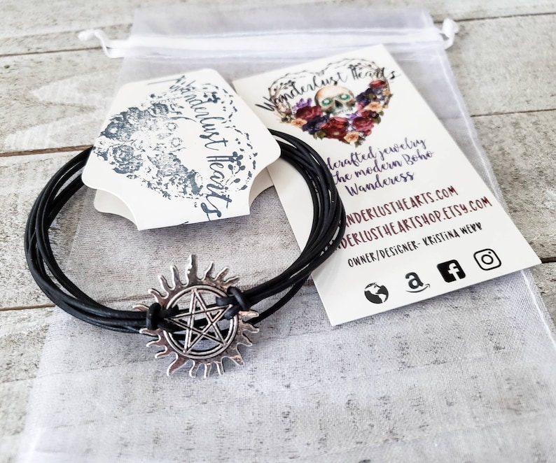 May include: Black leather bracelet with a silver charm featuring a pentagram inside a sunburst. The bracelet is packaged in a white organza bag with a business card that reads "Wanderlust Hearts" and includes the website address and social media links.