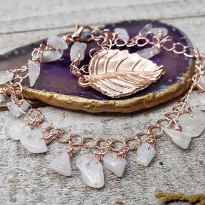 May include: A rose gold chain bracelet with a rose gold leaf charm and white moonstone beads hanging from the chain.