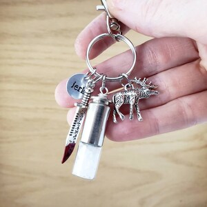 May include: Silver keychain with a small round charm that says "jerk", a silver knife charm, a silver vial charm, and a silver elk charm.