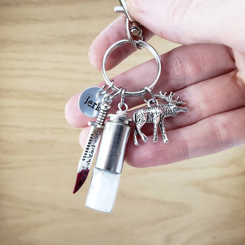 May include: Silver keychain with a small round charm that says "jerk", a silver knife charm, a silver vial charm, and a silver elk charm.
