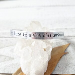 May include: A silver-colored cuff bracelet with the stamped text "I'm here to make shit awkward." The bracelet is displayed on a bed of clear quartz crystals.