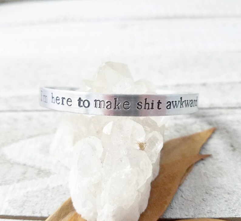 May include: A silver-colored cuff bracelet with the stamped text "I'm here to make shit awkward." The bracelet is displayed on a bed of clear quartz crystals.