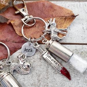 May include: Silver keychain with a small silver deer charm, a silver knife charm with a red handle, a silver bullet charm, a silver squirrel charm, and a silver charm with the word "jerk" on it.