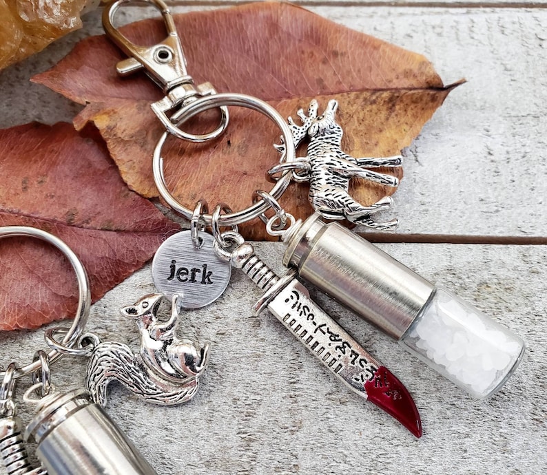 May include: Silver keychain with a small silver deer charm, a silver knife charm with a red handle, a silver bullet charm, a silver squirrel charm, and a silver charm with the word "jerk" on it.