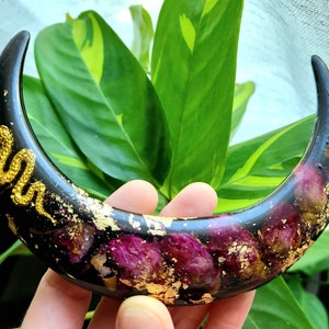 May include: A black crescent moon-shaped resin ornament with gold leaf and dried flowers embedded in the resin. A gold snake design is painted on the front of the ornament.